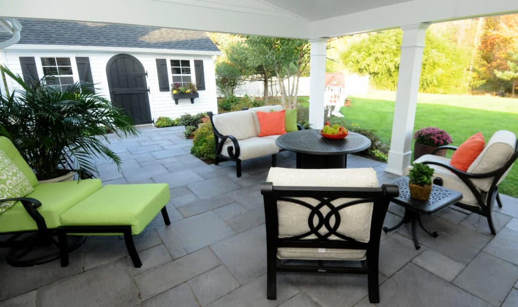Covered patio with cushioned chairs, a round table with a bowl of fruit, and colorful pillows. The space overlooks a garden and lawn with a white shed in the background. Lush plants and sunlight complete the inviting scene.