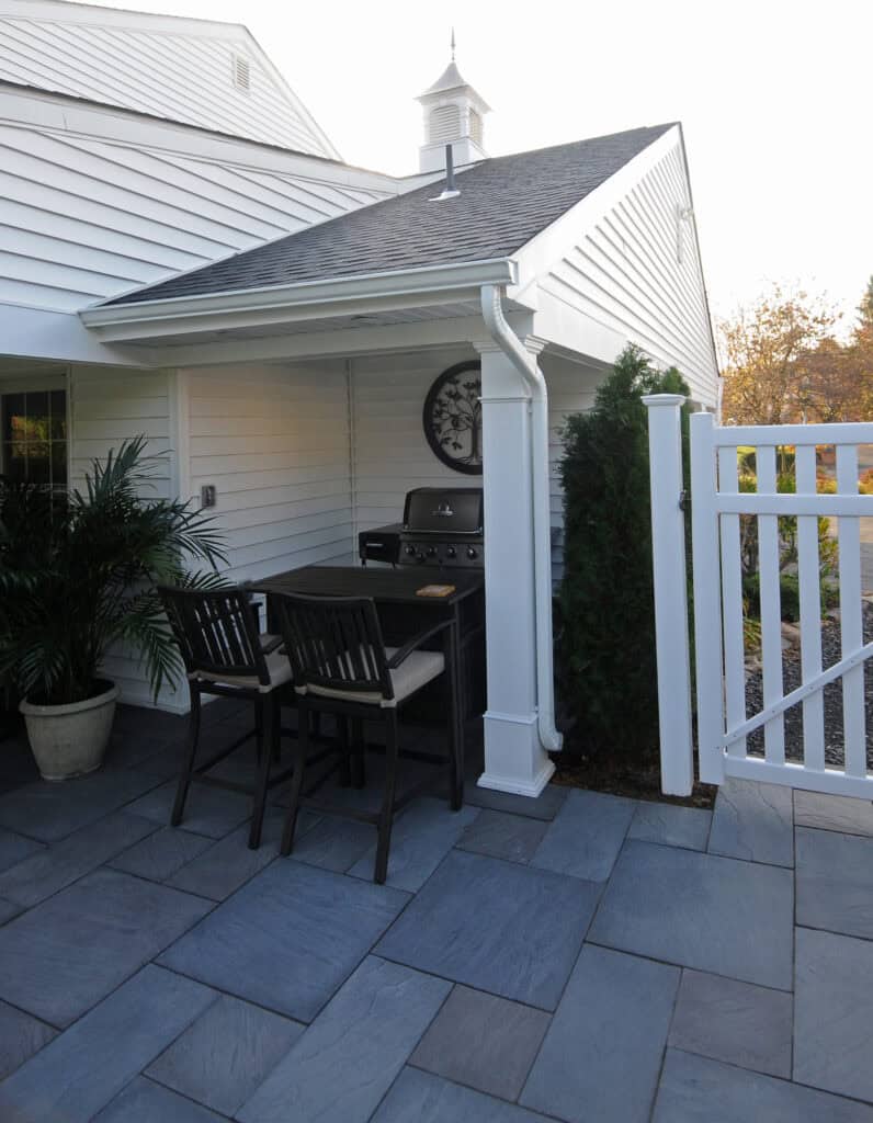 A small outdoor patio with slate tile flooring features a grill, a wall clock, two barstools at a counter, a potted plant, and a white fence next to a house with white siding.
