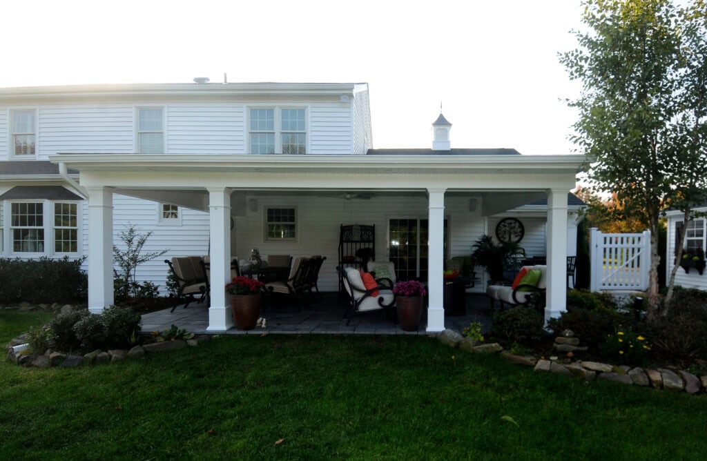 A white house with a covered patio featuring outdoor seating, potted plants, and cushions. The patio overlooks a green lawn bordered by stone edging and landscaping. The scene is bright and inviting.