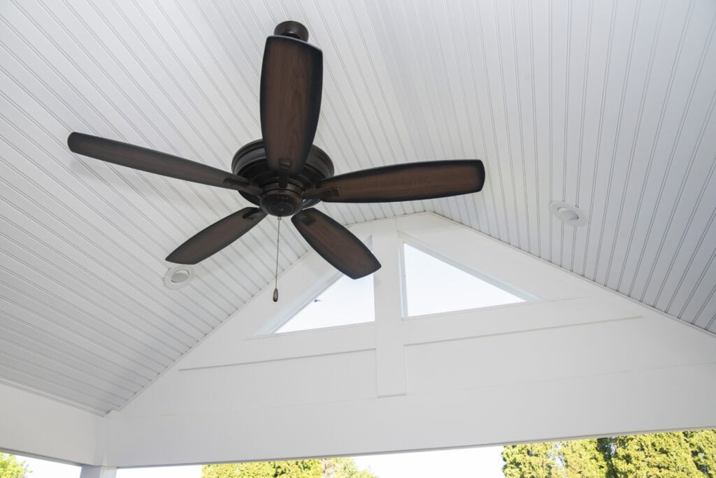 Brown ceiling fan with five blades mounted on a white, slanted, paneled ceiling; triangular windows and some greenery are visible through the open space below the ceiling.