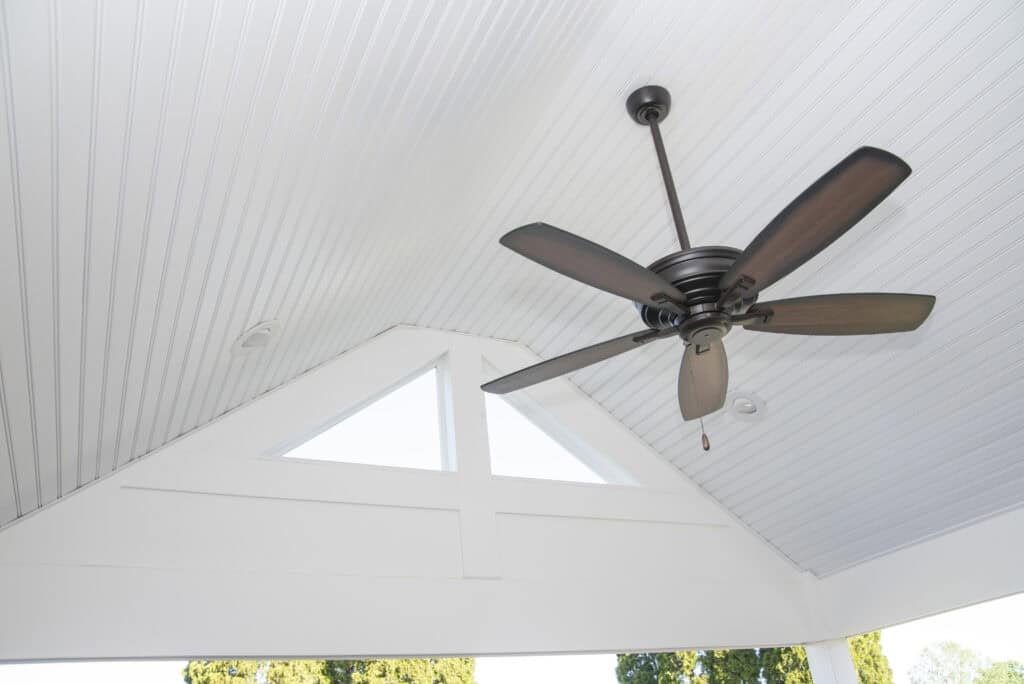 A black ceiling fan with five blades hangs from a white, vaulted, beadboard ceiling with triangular windows letting in natural light. Green trees are visible outside.