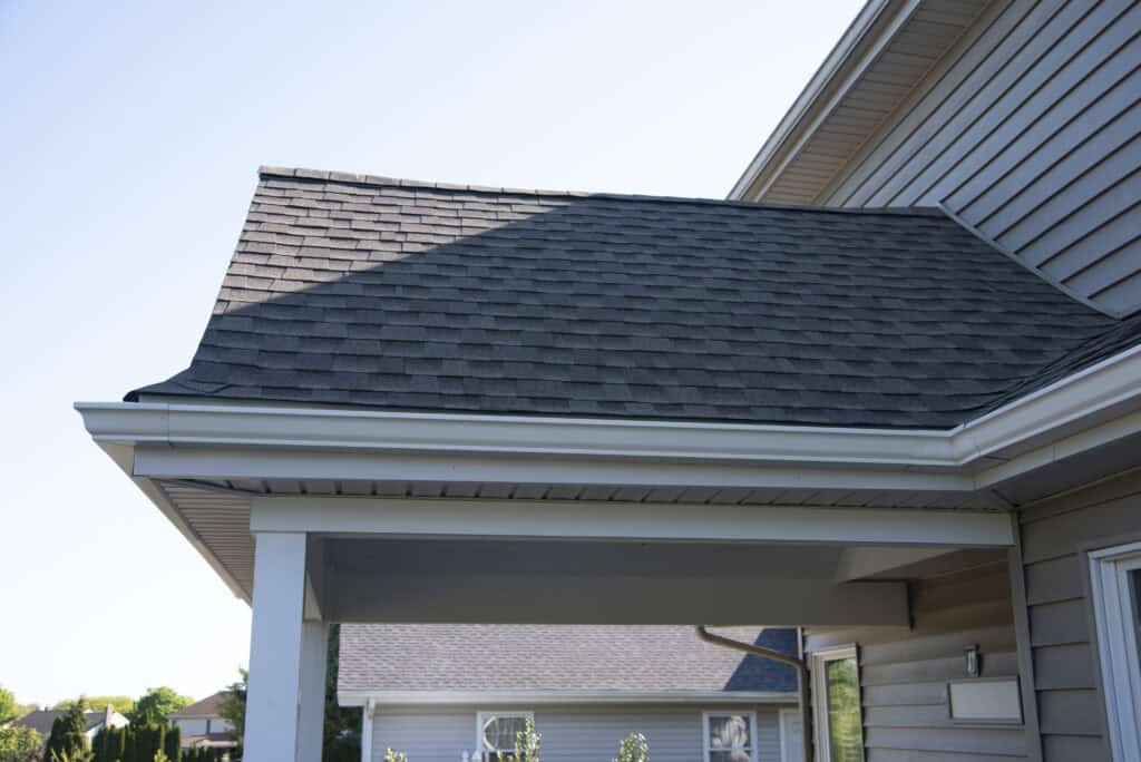 A close-up view of a house roof with dark asphalt shingles, white gutters, and beige siding, taken on a clear, sunny day. Part of another house and green plants are visible in the background.
