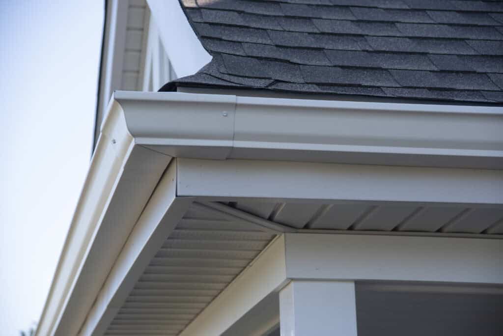 Close-up of a house corner showing gray roof shingles, white rain gutters, downspout, and soffit with vented panels under the eaves.