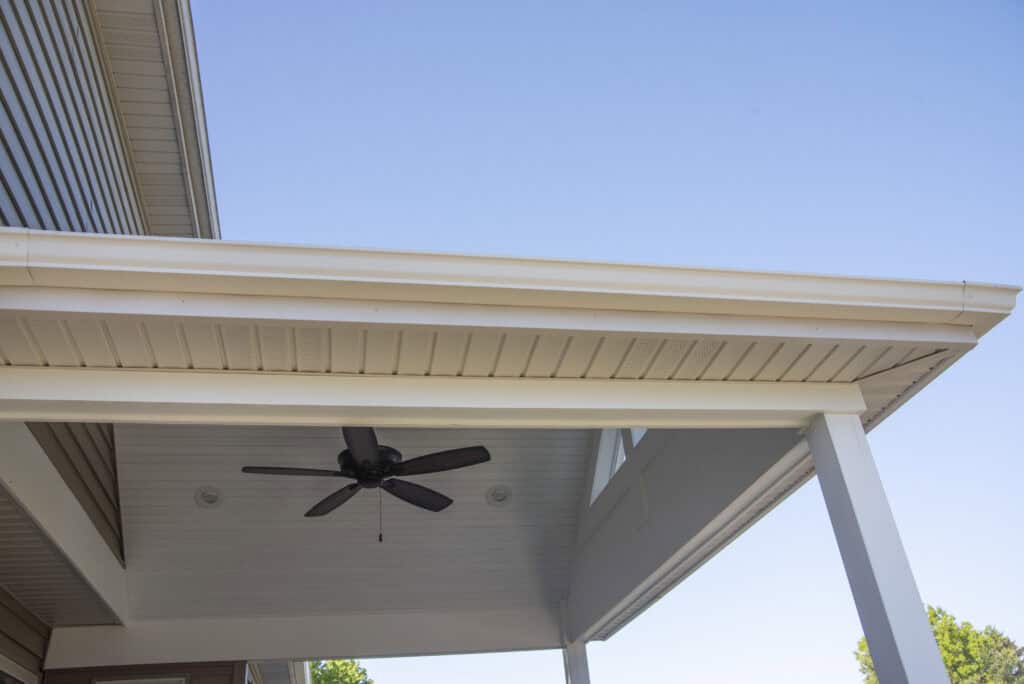 A close-up view of a covered patio with a white roof and support beams, featuring a black ceiling fan underneath and a clear blue sky in the background.