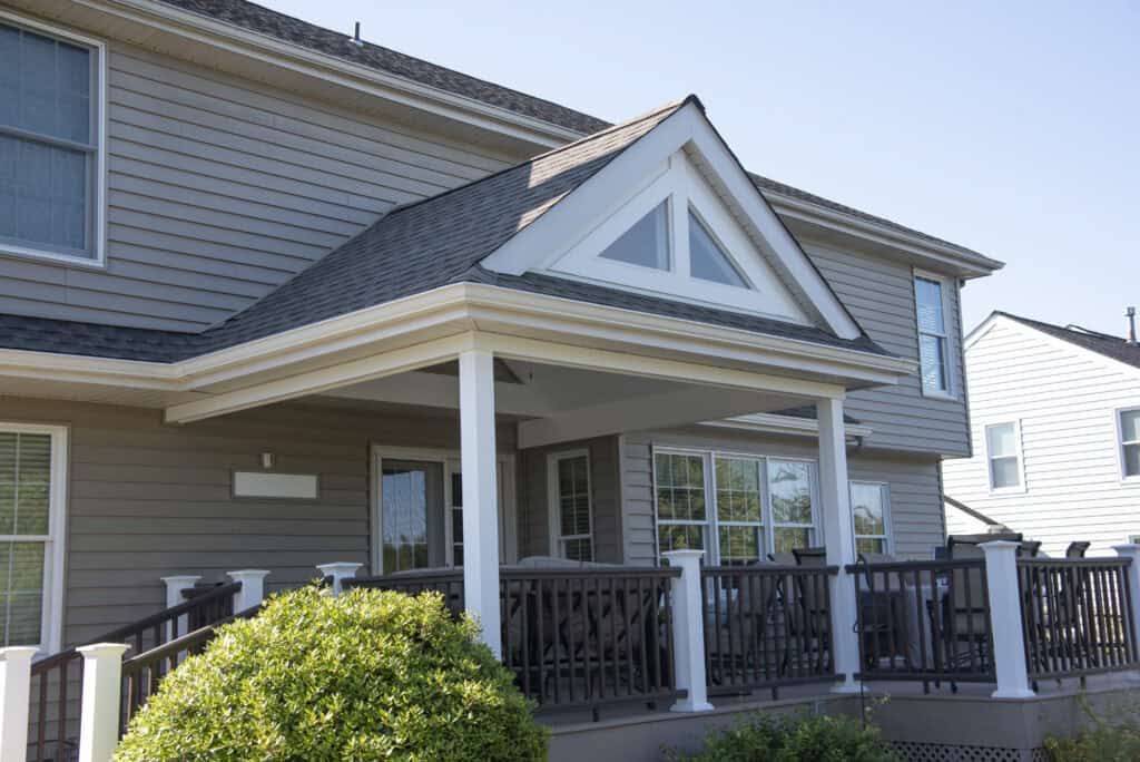 A two-story beige house with white trim features a covered porch with dark railings and several windows. There are green bushes in the foreground under a clear blue sky.