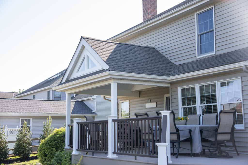 A modern suburban house with beige siding, a covered porch, white trim, and outdoor furniture including a table and chairs. There are some bushes and landscaped greenery around the porch.
