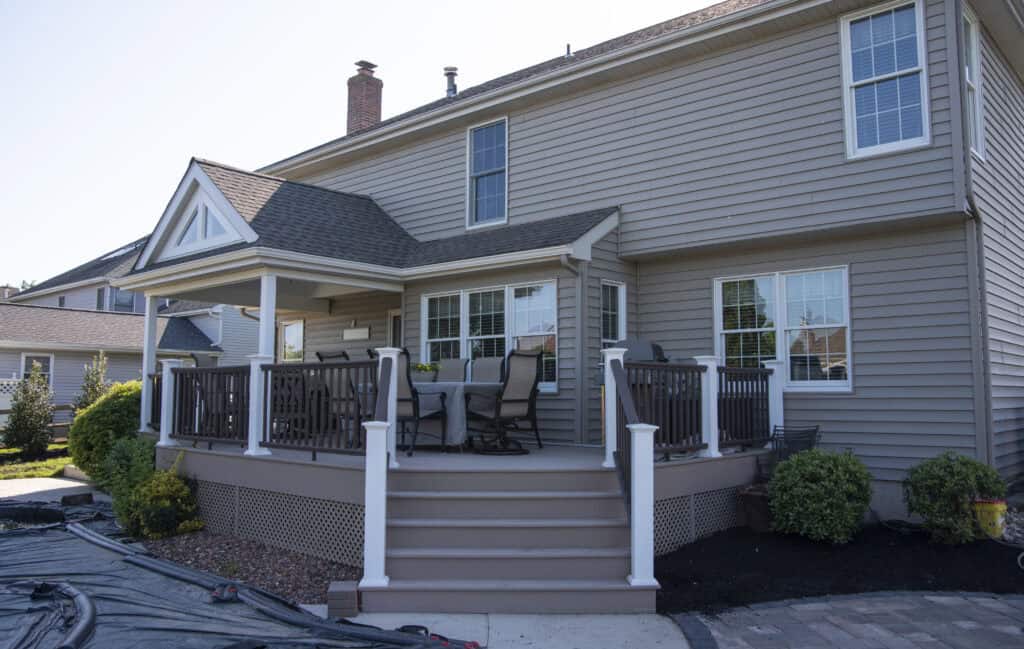 A two-story beige house with a covered porch and wooden deck featuring patio furniture. The deck has stairs with white railings, and the landscaping includes bushes and mulch around the house.