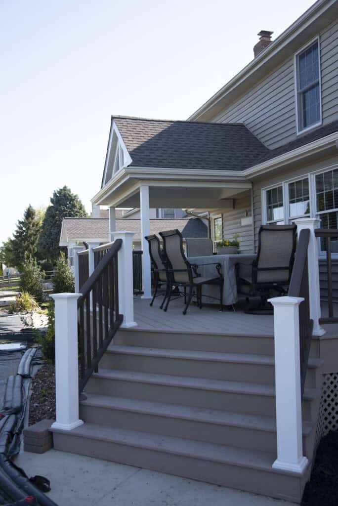 A raised wooden deck with stairs, white railings, and a covered seating area with four chairs and a table next to a beige house on a sunny day.
