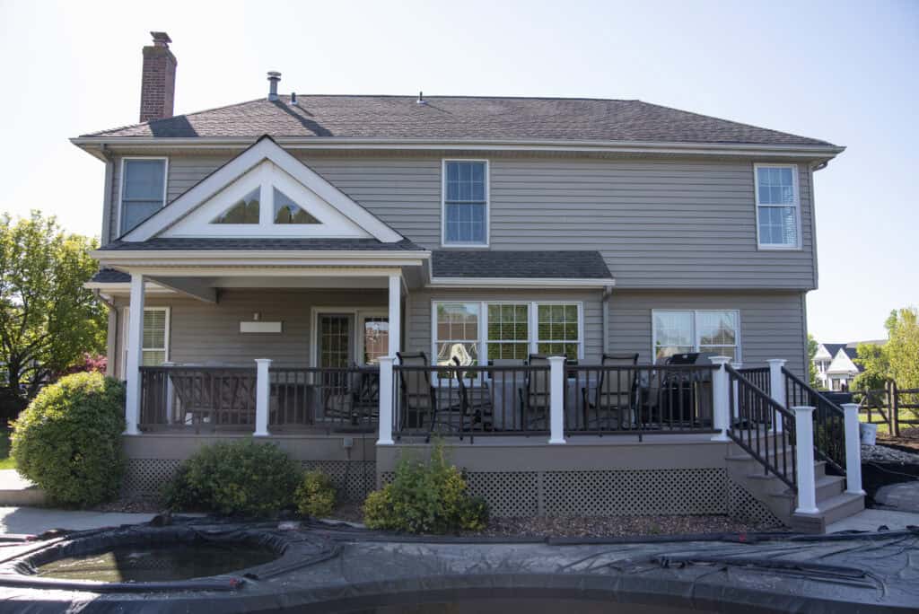 A two-story suburban house with beige siding, a covered back porch with white railings, patio furniture, and landscaped bushes beside a pool with a cover on it.