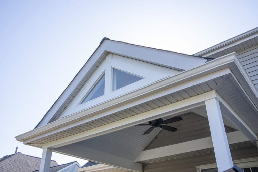 The image shows the roof and covered porch of a modern house with white trim, beige siding, triangular windows, and a ceiling fan beneath the porch roof against a clear blue sky.