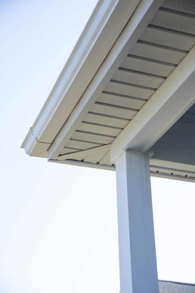 Close-up view of a house roof corner showing beige soffit, fascia, and white rain gutter above a supporting white column, with a clear blue sky in the background.
