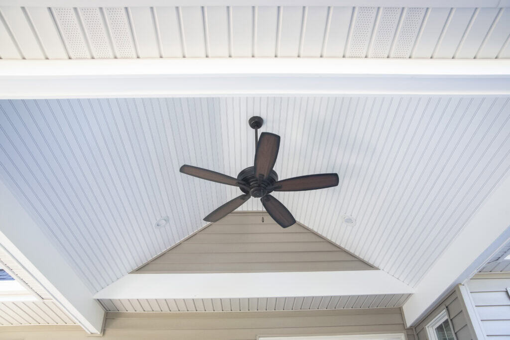 A black ceiling fan with five blades hangs from the center of a white, vaulted, beadboard ceiling in a covered outdoor patio area. The ceiling features angled beams and soft lighting.