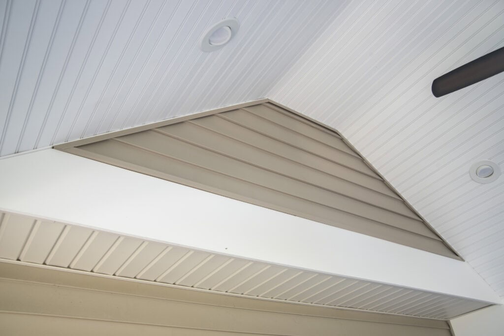 A close-up view of a vaulted ceiling with white beadboard paneling, recessed lights, and a triangular section of beige horizontal siding.