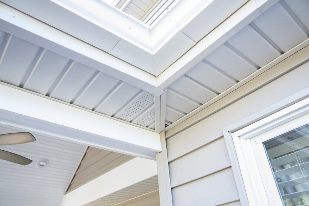 Close-up view of the corner of a house exterior showing white soffit, fascia, and beige vinyl siding, with part of a ceiling fan and window with blinds visible.