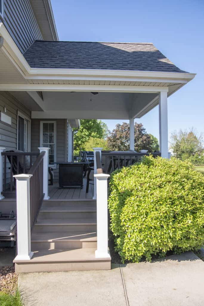 A small porch with steps, white railing, and a shaded roof attached to a house. There is patio furniture on the porch and a neatly trimmed green bush beside the steps under a clear blue sky.