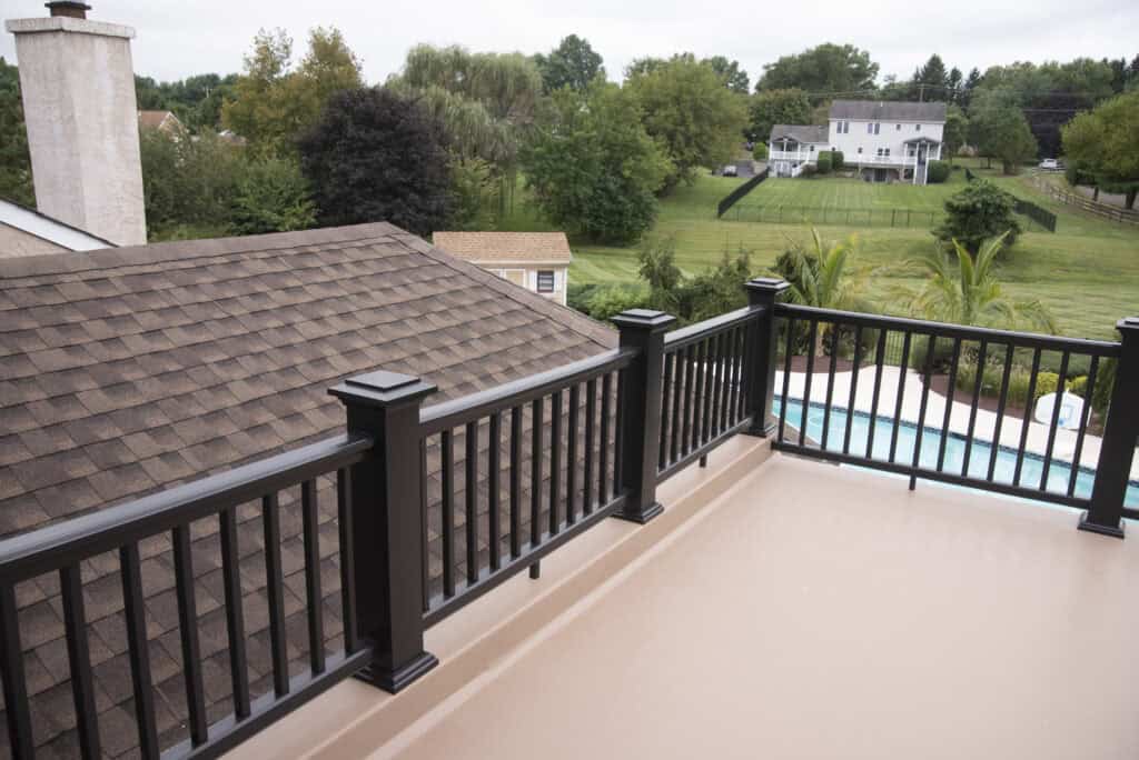 A view from a balcony with a brown floor and black railing, overlooking a shingled roof, green lawn, trees, a swimming pool, and houses in the distance.