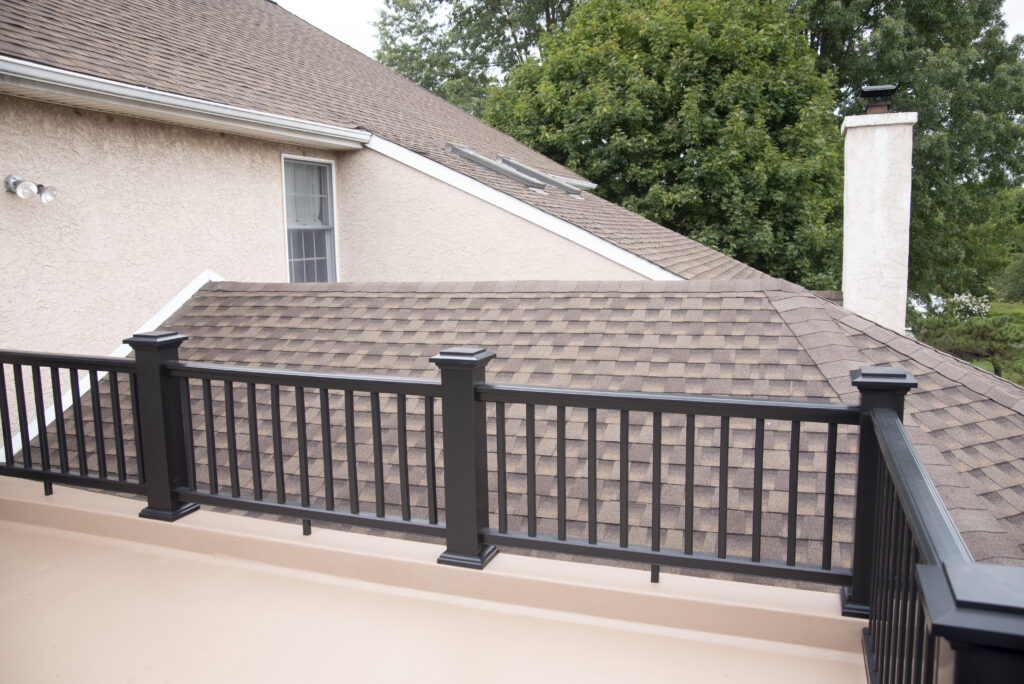 A view from a balcony with a black metal railing, overlooking a house with brown shingle roofs and beige walls. Green trees are in the background.