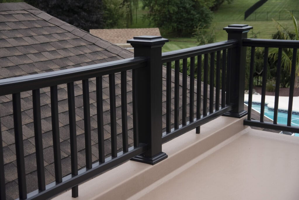 A close-up view of a black metal railing on a balcony overlooking a brown shingle roof, green trees, and a pool in the background. The scene appears to be part of a residential area.
