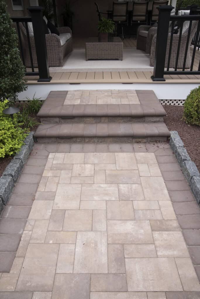 A paved walkway with stone tiles leads up two steps to a covered patio area with wicker furniture and a table, bordered by plants and shrubs.