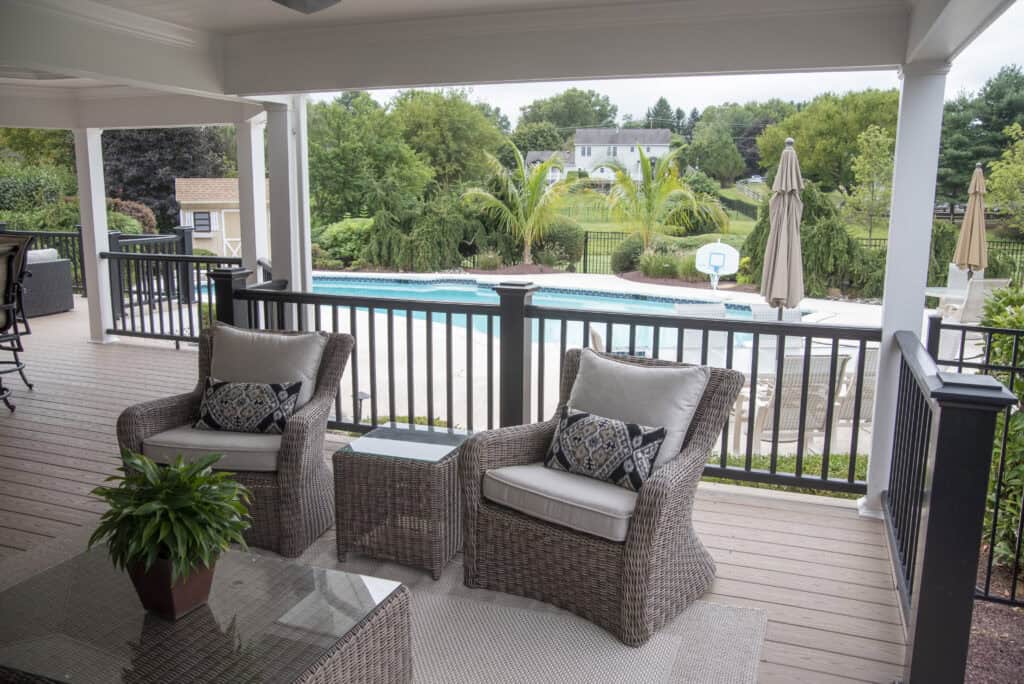 Two wicker chairs with cushions and a small table sit on a covered wooden deck overlooking a backyard with a swimming pool, lounge chairs, umbrellas, and green trees in the background.