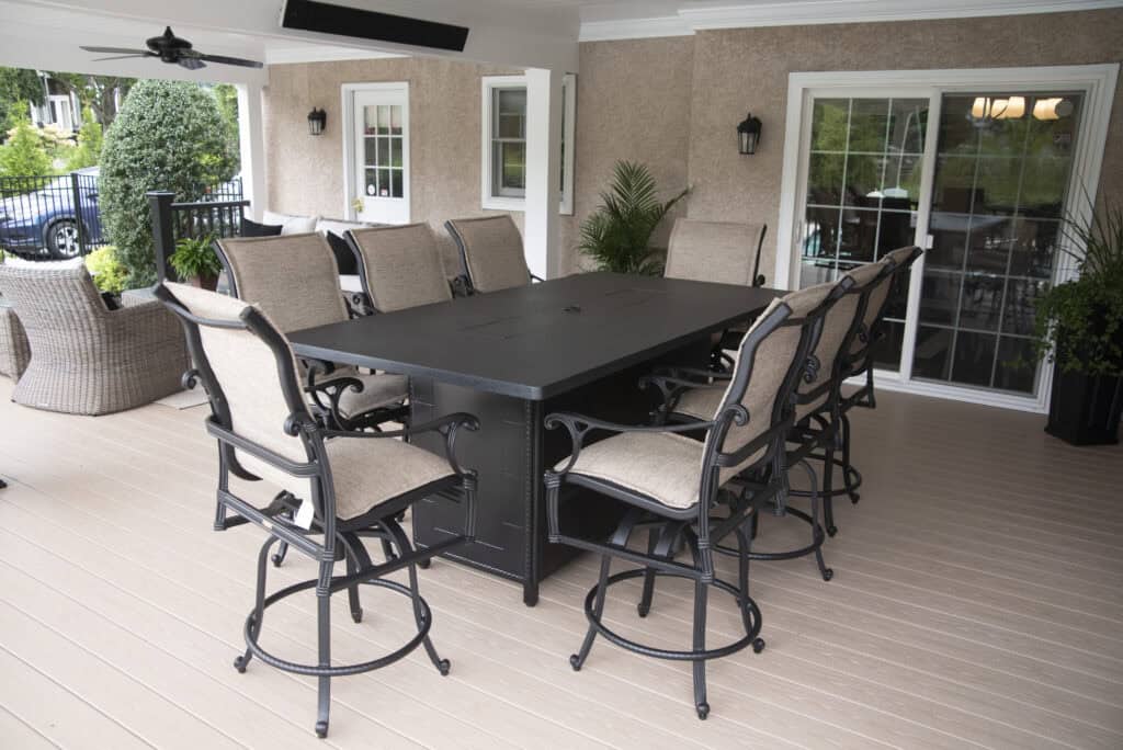 A large black rectangular outdoor dining table surrounded by ten cushioned chairs sits on a covered patio with beige flooring. Potted plants and sliding glass doors are visible in the background.
