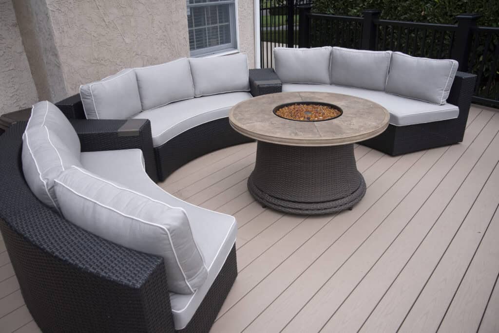 Curved outdoor wicker sofas with light gray cushions surround a round fire pit table on a light-colored deck next to a house. Black railing and greenery are in the background.