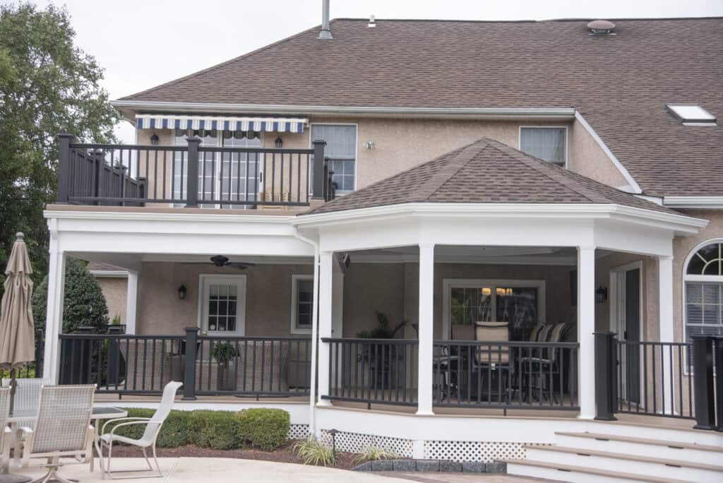 A two-story house with a large covered porch and balcony, white pillars, black railings, outdoor furniture, and a striped awning on the upper level. Shrubs and chairs are arranged around the porch area.