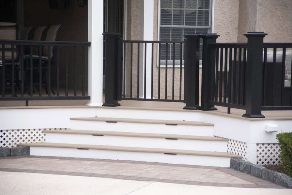 Four wide beige steps with black railings lead up to a porch with outdoor seating. The porch has white trim, a lattice skirting, and is attached to a house with light brown walls and windows.