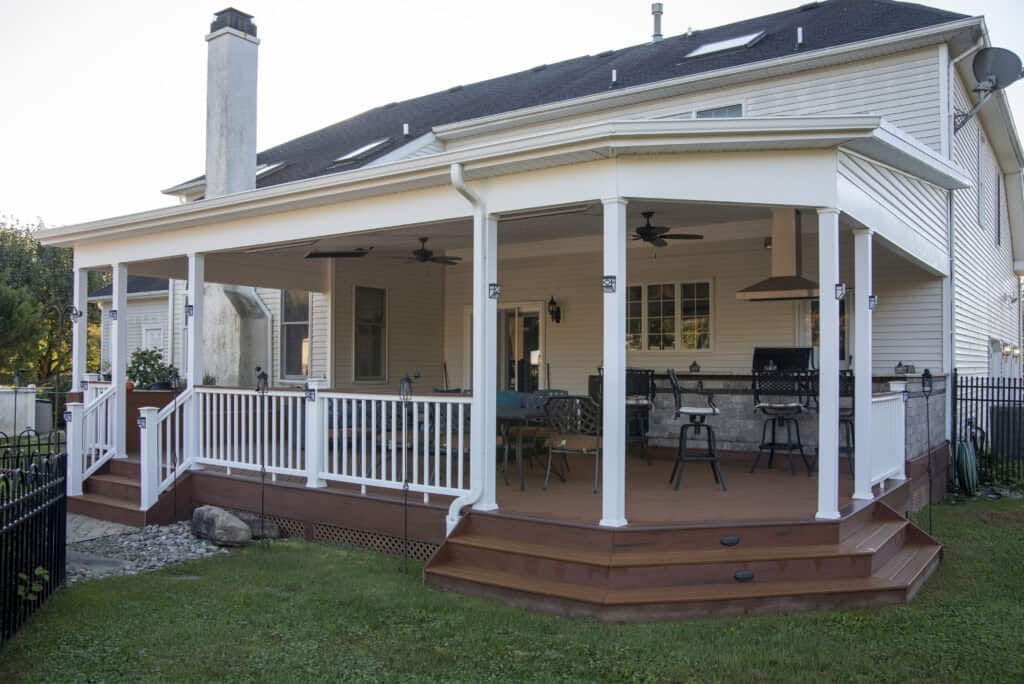 A spacious covered backyard deck attached to a house, featuring white railings, ceiling fans, a grill, bar seating, and patio furniture, with steps leading down to a green lawn.
