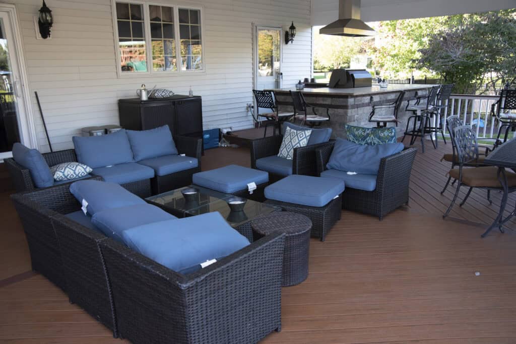 Outdoor patio with dark wicker furniture and blue cushions, arranged around a glass coffee table. In the background, there is a bar area with stools, a grill, and a countertop under a covered roof.
