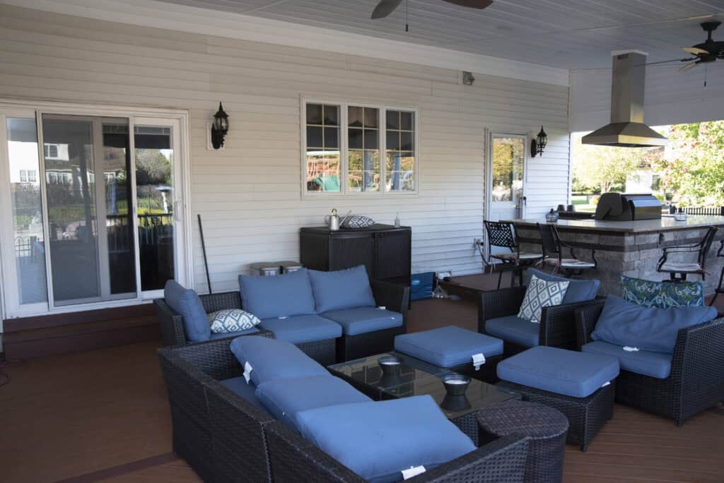 A covered patio features a wicker outdoor seating set with blue cushions, a glass coffee table, a grill area with barstools, and sliding glass doors leading inside. The space is well-lit and neatly arranged.