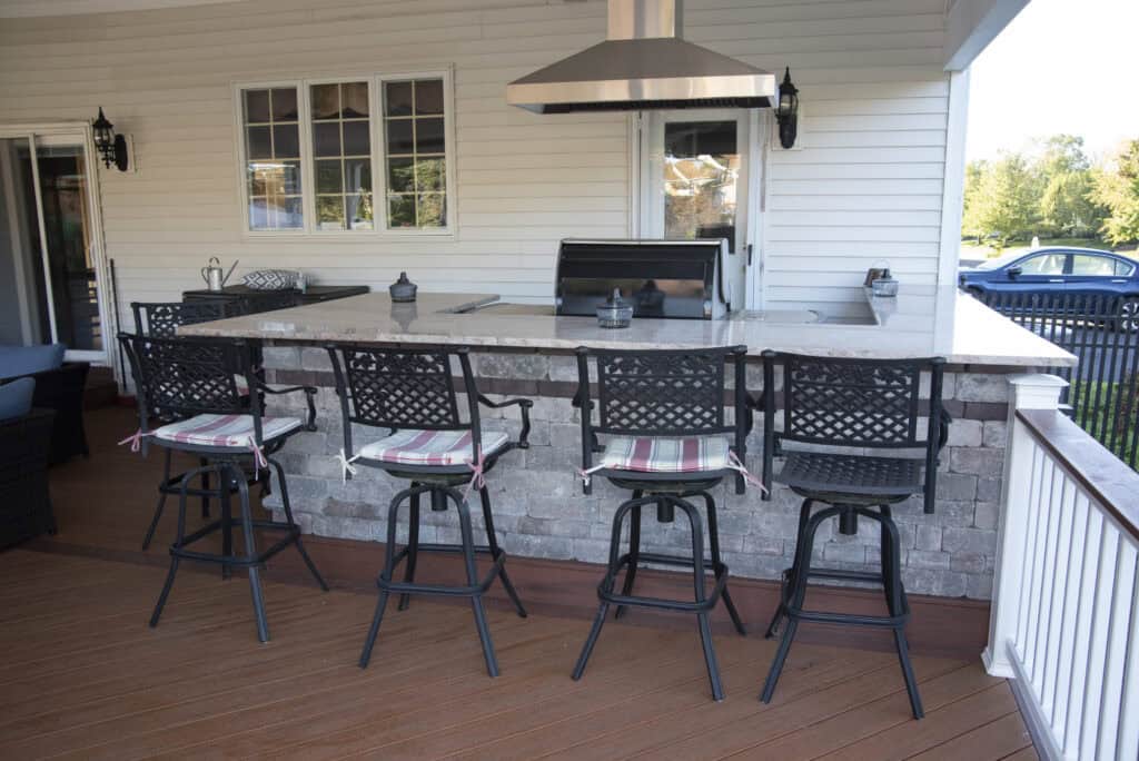 Outdoor kitchen with a stone island, granite countertop, four black metal barstools with striped cushions, grill, and chimney vent on a wooden deck. White siding and windows are in the background.