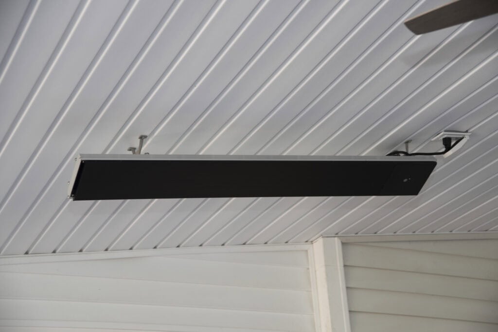 A black rectangular ceiling-mounted heater or infrared heater is attached to a white paneled ceiling in a covered outdoor area with white walls.