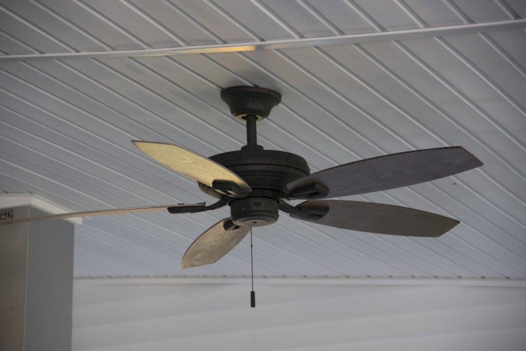 A dark-colored ceiling fan with five blades and a pull chain hangs from a white paneled ceiling. The fan is off, and the background is a light, indoor setting.