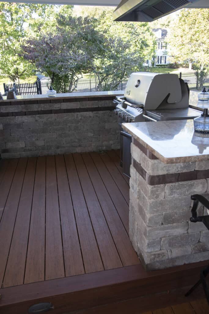 Outdoor patio with a built-in stone grill, stainless steel barbecue, and a marble countertop. Wooden deck flooring and trees with green leaves are visible in the background beyond a black fence.