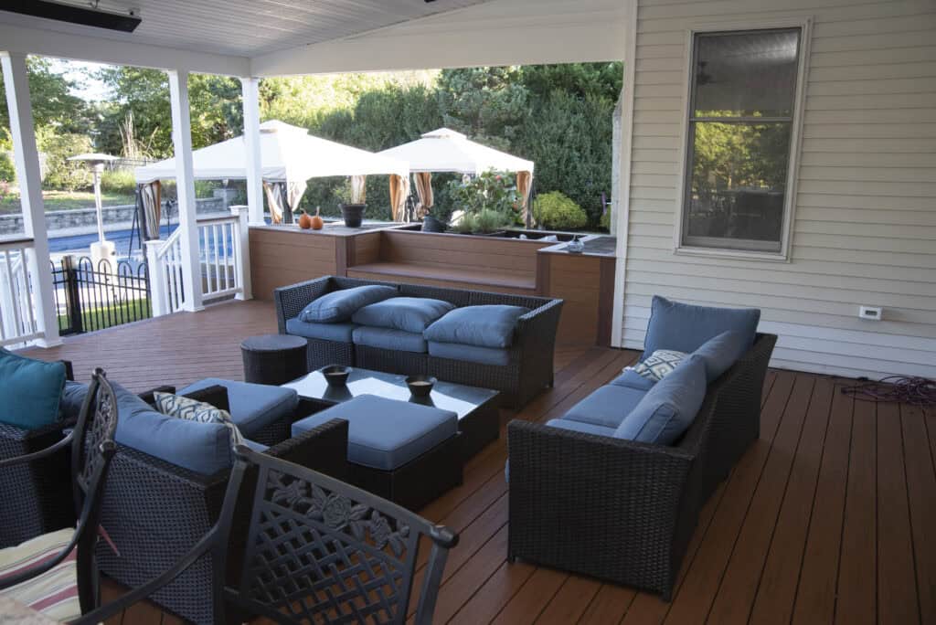 Outdoor patio with wicker furniture, blue cushions, a glass coffee table, and a wooden deck. Large umbrellas and a pool are visible in the background, surrounded by greenery and a privacy fence.