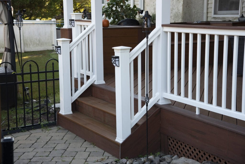 Wooden porch steps with white railings lead up to a deck with brown flooring. A black metal fence and stone path are in the foreground, with garden decor and plants visible around the porch.