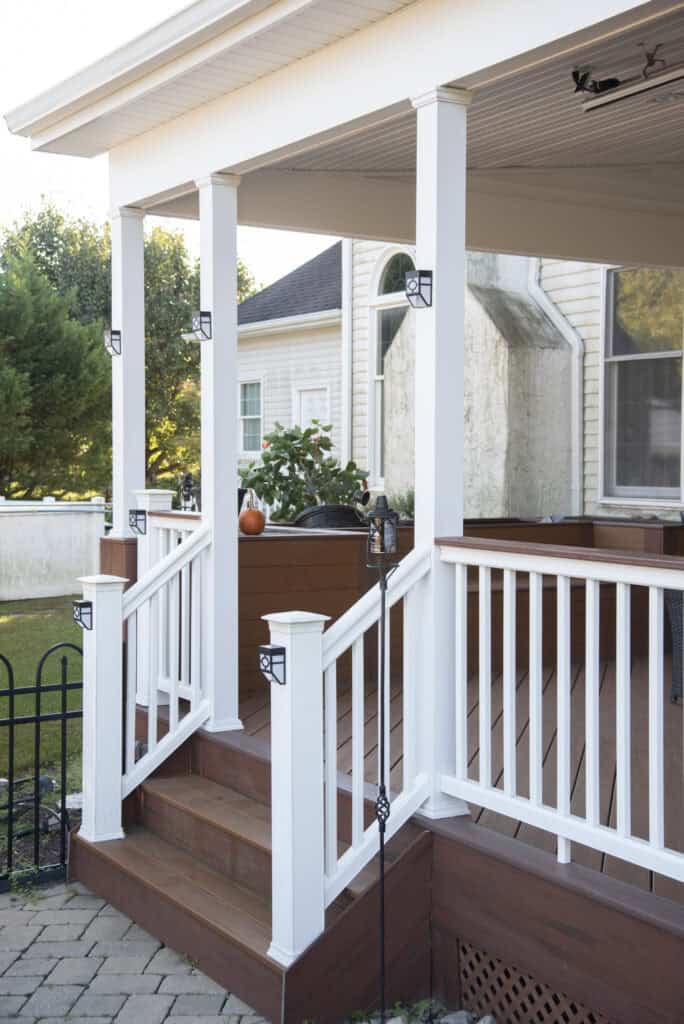 A wooden porch with white railings and posts, brown steps, and a small pumpkin on a planter near the house. The porch is attached to a light-colored house with trees in the background.