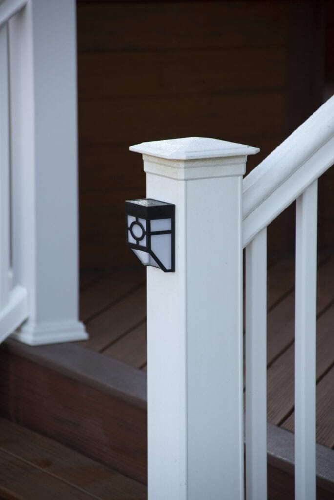 A white porch railing post with a decorative black and white light fixture attached, set against brown wooden steps and siding in the background.
