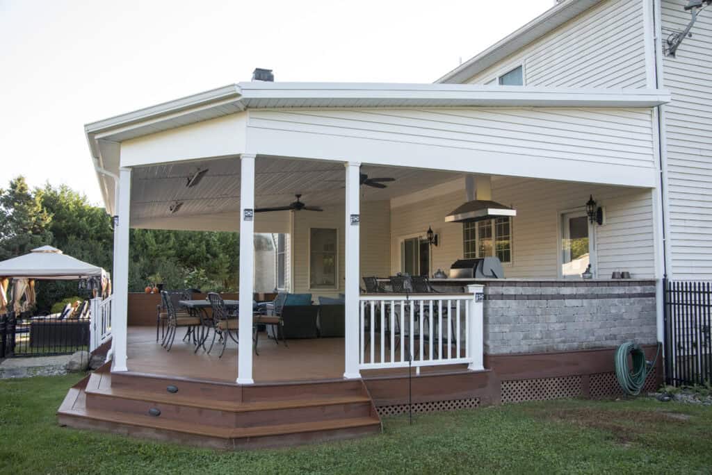 A covered backyard patio attached to a house with white siding, featuring outdoor seating, a dining table, ceiling fans, a grill, and wooden steps leading to a grass lawn. A garden hose is coiled by the patio.