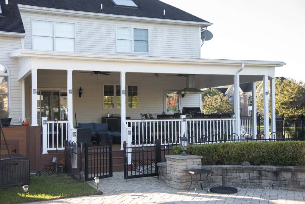 A two-story house with a covered back porch featuring outdoor furniture, ceiling fans, and a fenced area. The porch overlooks a stone patio with a fire pit and landscaped greenery.