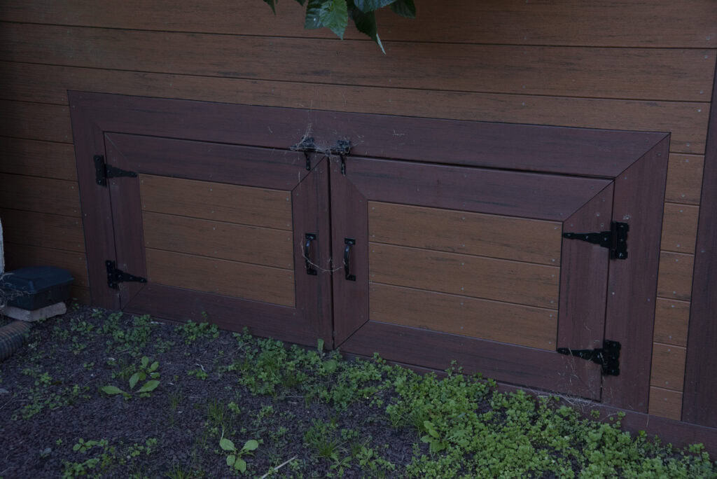 A pair of brown wooden doors with black metal hinges and handles set into the lower part of a wall, surrounded by green plants and soil.