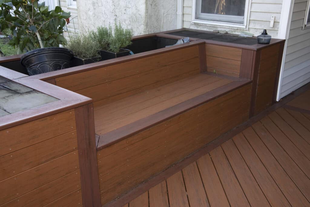 A wooden outdoor bench with built-in planters on both sides, placed on a wooden deck next to a house. Several potted plants and herbs are arranged in the planters.
