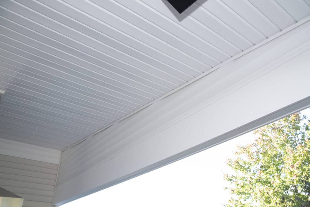 White paneled ceiling with clean lines and a large beam, seen from below. Part of a tree with green leaves appears at the lower right edge against a bright sky.
