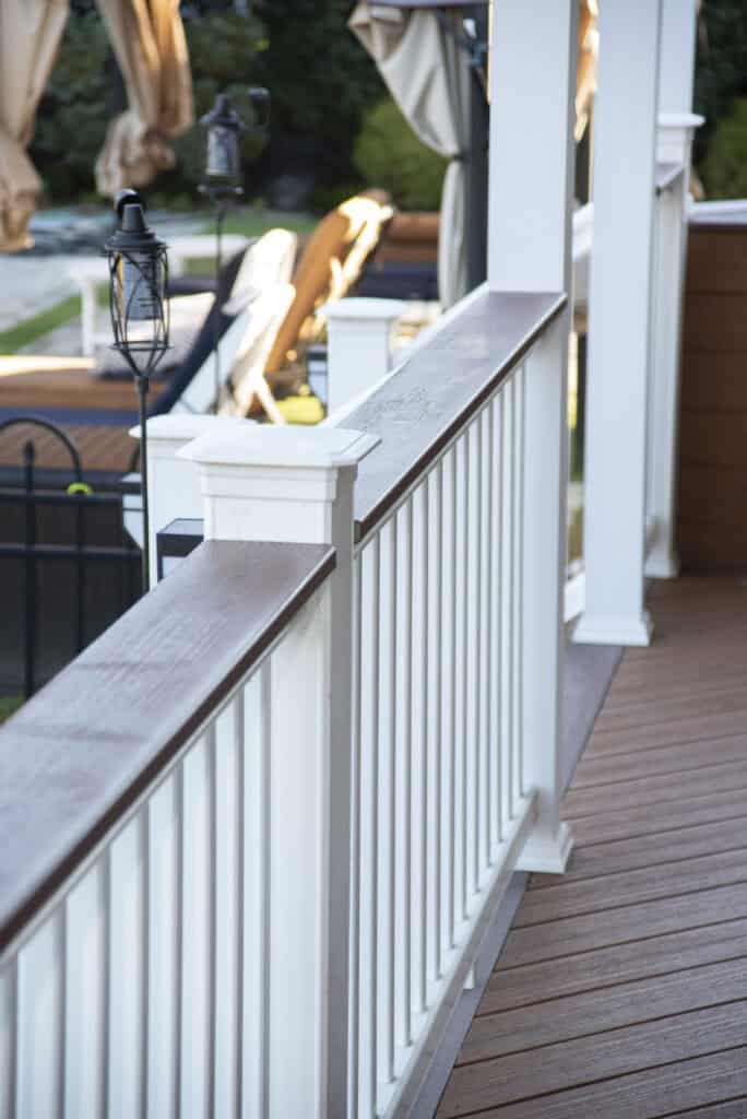 White railing with a brown wooden top on a deck with brown flooring; outdoor furniture and a garden area with chairs and umbrellas are visible in the background.