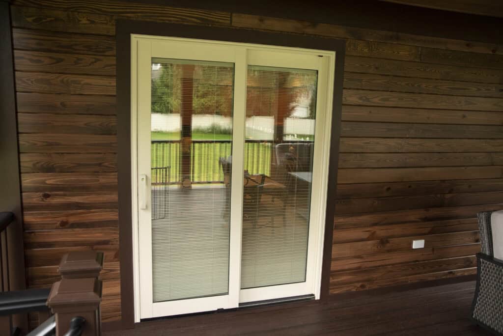 White-framed sliding glass door with built-in blinds, set in a wooden wall on a porch. The door opens to a covered deck with a view of a green yard and trees in the background.
