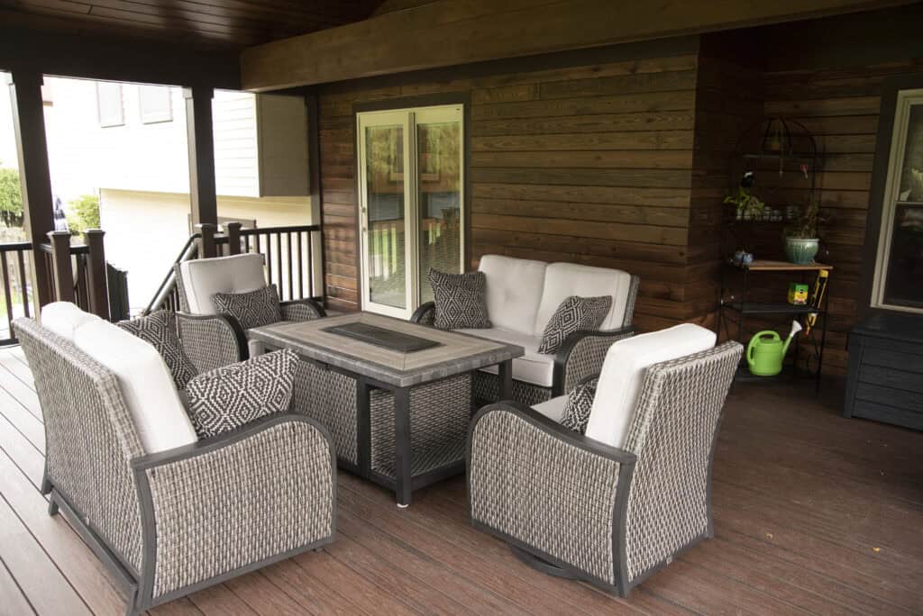 A covered patio with wicker patio furniture, including a sofa, four chairs, and a rectangular fire pit table. The wood-paneled walls and deck create a cozy outdoor seating area with patterned pillows and potted plants.