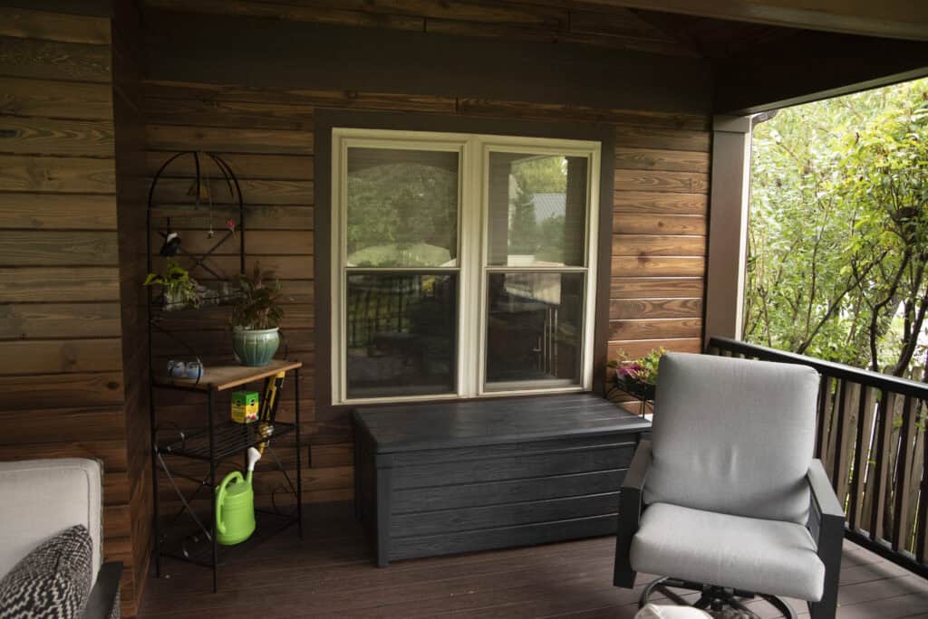 A cozy wooden porch features a window, a black storage bench, a metal shelf with plants and gardening tools, a gray cushioned chair, and green trees visible outside the railing.