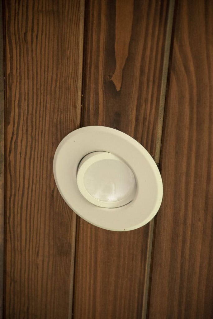 A recessed round white light fixture is mounted on a wooden ceiling with a natural finish and visible wood grain.