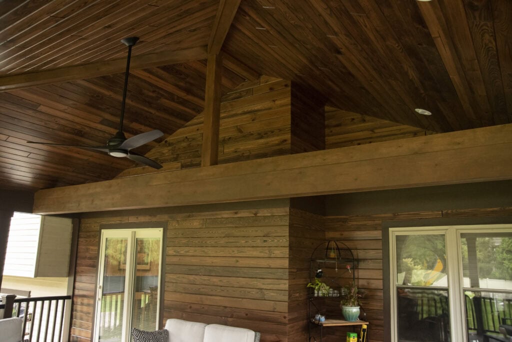 A covered patio with a wooden ceiling and walls, a ceiling fan, sliding glass doors, and some outdoor furniture and decorations. Natural light comes in from windows and doors.